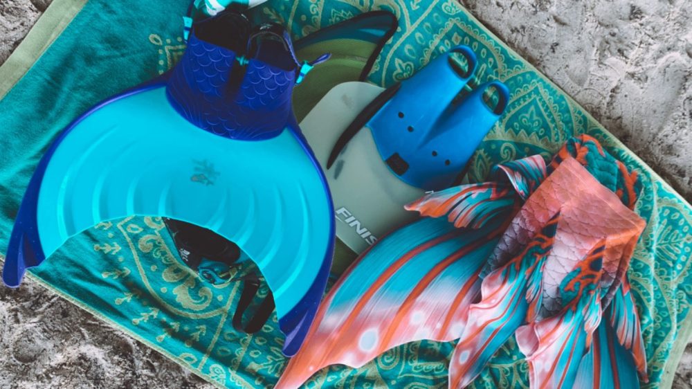 Mermaid tail swimsuits and fins on a green beach towel on sand.