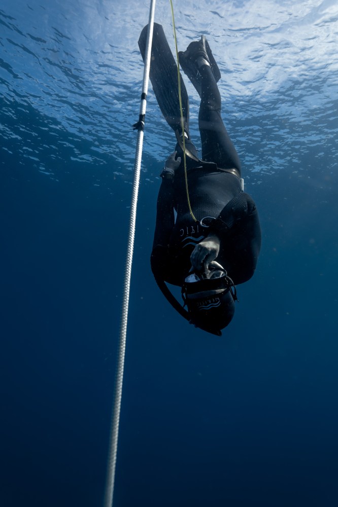 A diver in a wetsuit descends underwater alongside a rope, with sunlight filtering through the water.