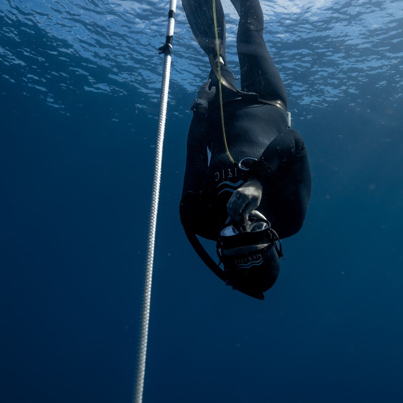 A diver in a wetsuit descends underwater alongside a rope, with sunlight filtering through the water.