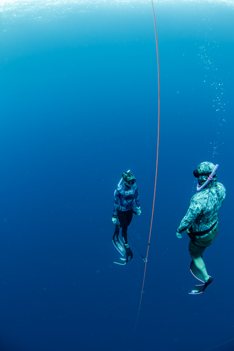 Two divers in wetsuits floating underwater beside a guide rope in deep blue ocean.