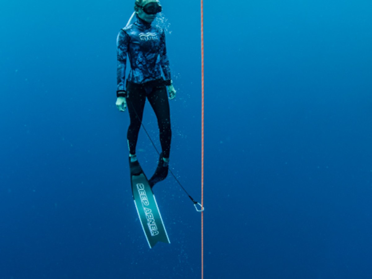 Diver with fins and snorkel floating vertically beside a rope in deep blue water.