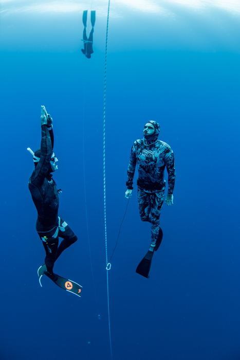Two divers in wetsuits underwater, one swimming upwards, one holding a rope, blue ocean backdrop.