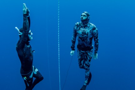 Two divers in wetsuits underwater, one swimming upwards, one holding a rope, blue ocean backdrop.