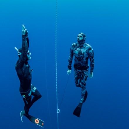 Two divers in wetsuits underwater, one swimming upwards, one holding a rope, blue ocean backdrop.