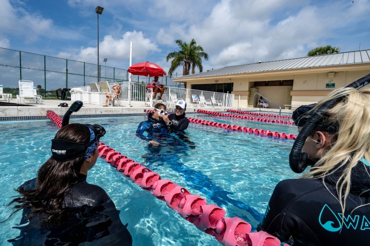 Group of people in wetsuits and snorkels having a lesson in an outdoor pool.