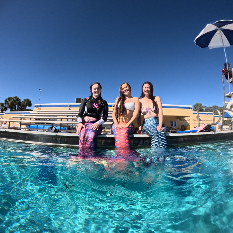 Three women wearing mermaid tails sitting by a pool, bright day, clear sky.