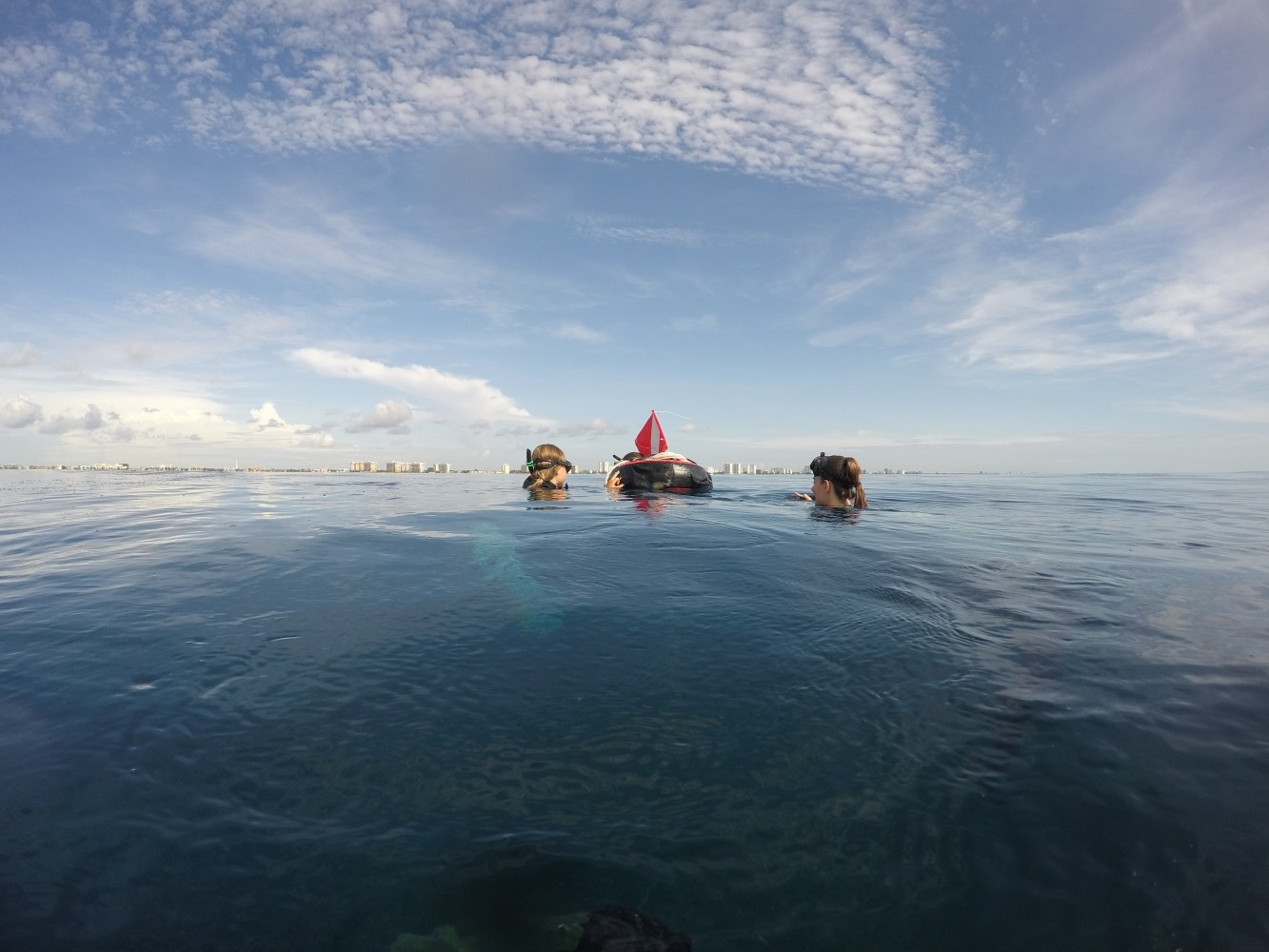 Two snorkelers surface near a red dive flag in calm ocean waters under a partly cloudy sky.