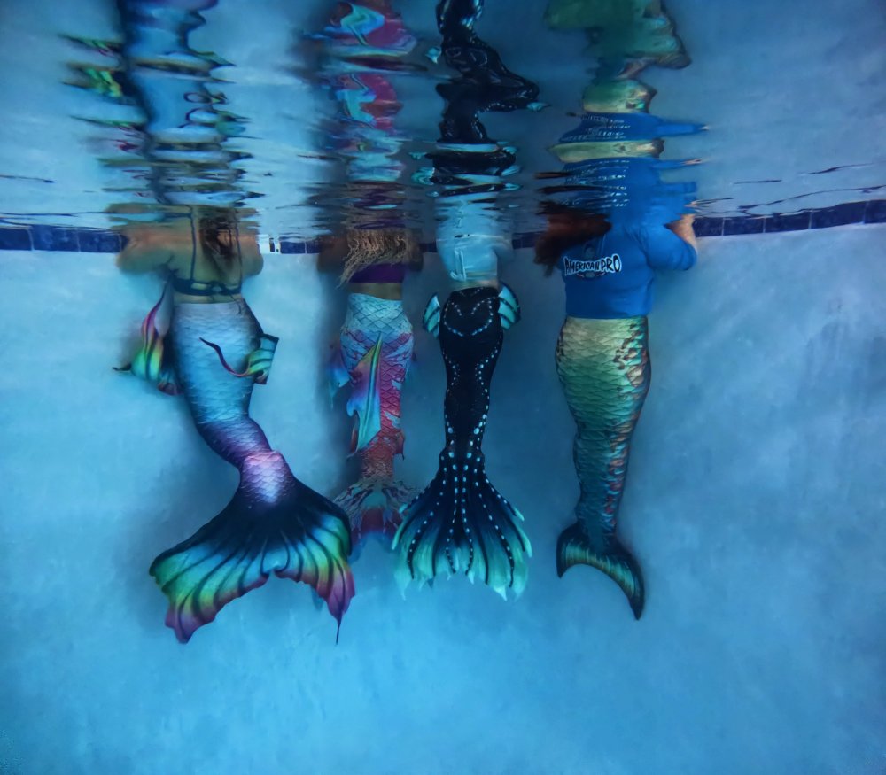 Four people underwater wearing colorful mermaid tails in a swimming pool.