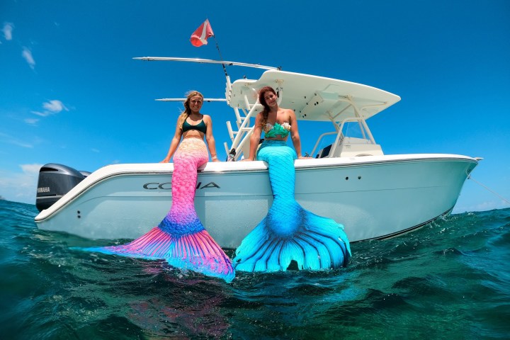 Two people in mermaid costumes sit on a boat in the ocean under a clear blue sky.