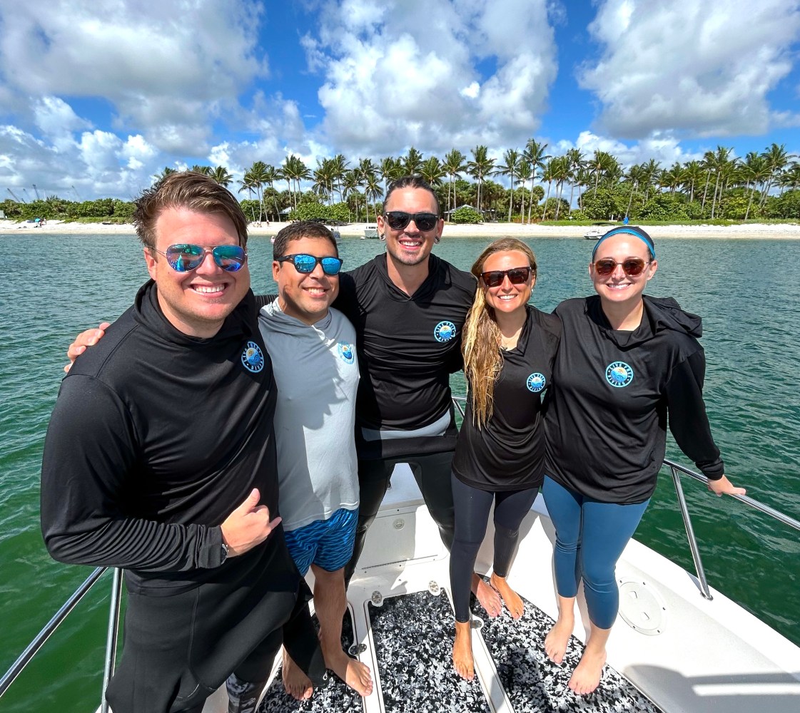 Five people smiling on a boat, wearing sunglasses, with a tropical island in the background.