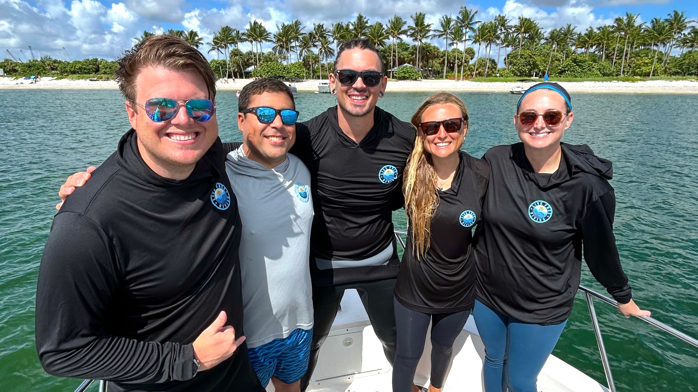 Five people smiling on a boat, wearing sunglasses, with a tropical island in the background.