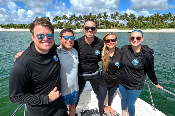 Five people smiling on a boat, wearing sunglasses, with a tropical island in the background.