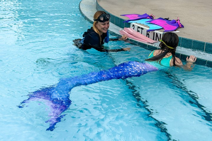 Person teaching girl in mermaid tail at swimming pool edge, colorful fins on deck.