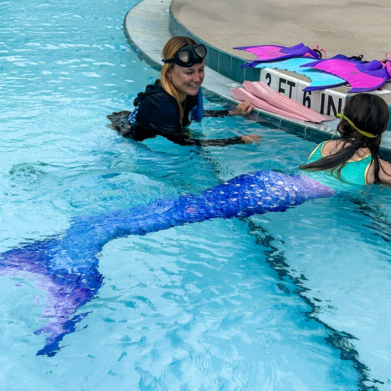 Person teaching girl in mermaid tail at swimming pool edge, colorful fins on deck.