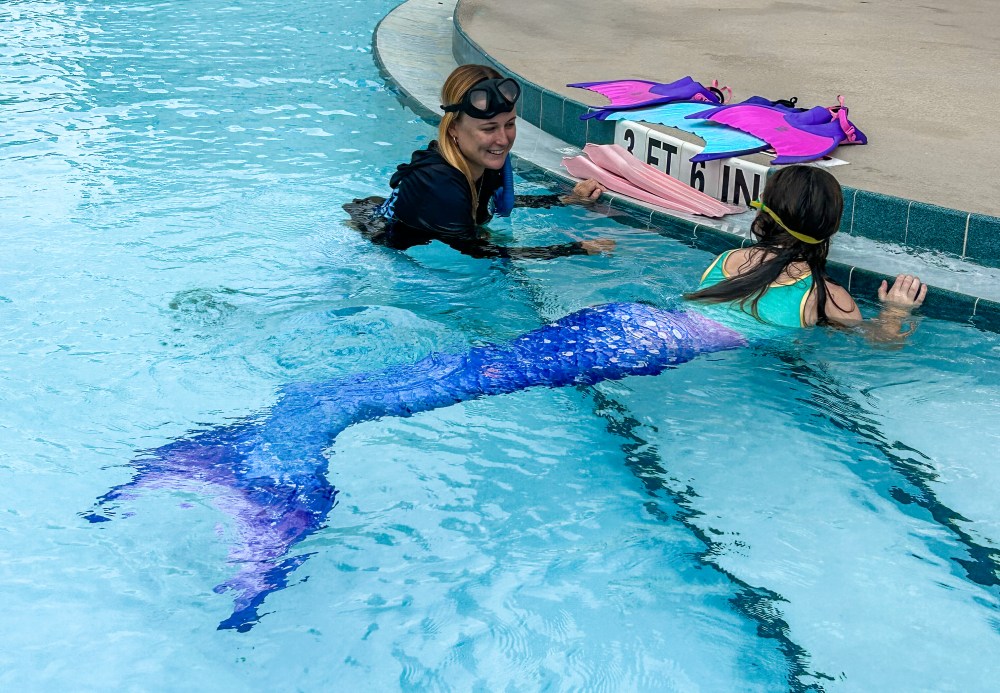 Person teaching girl in mermaid tail at swimming pool edge, colorful fins on deck.