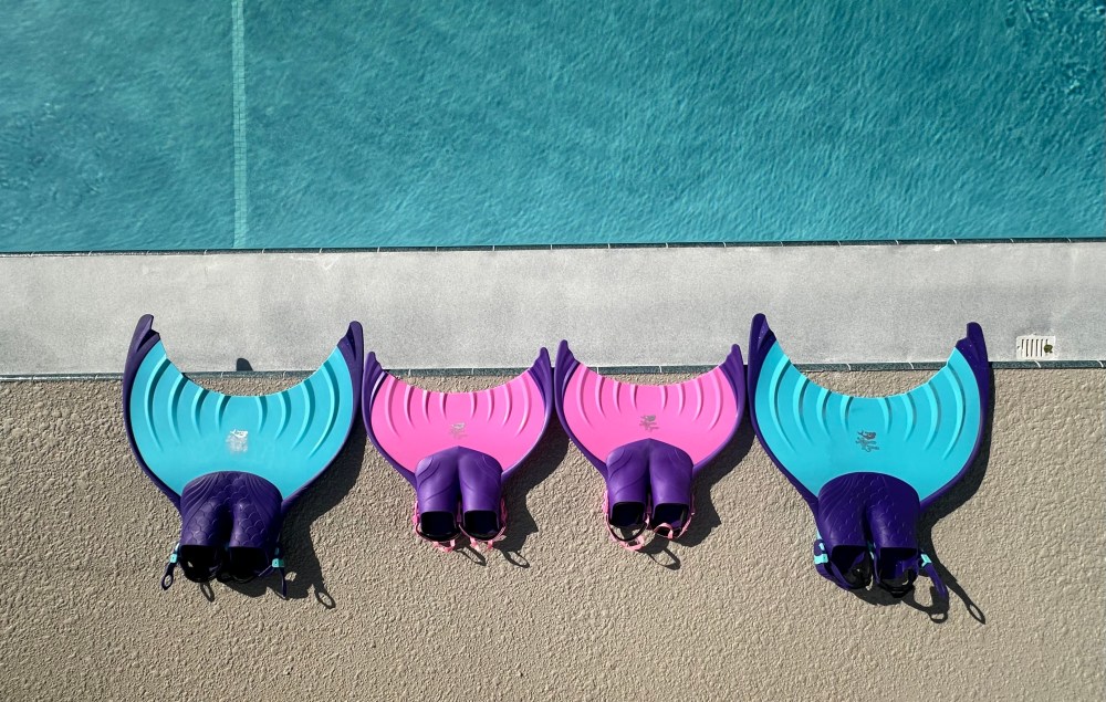Four mermaid tails in blue and pink on poolside near clear water.