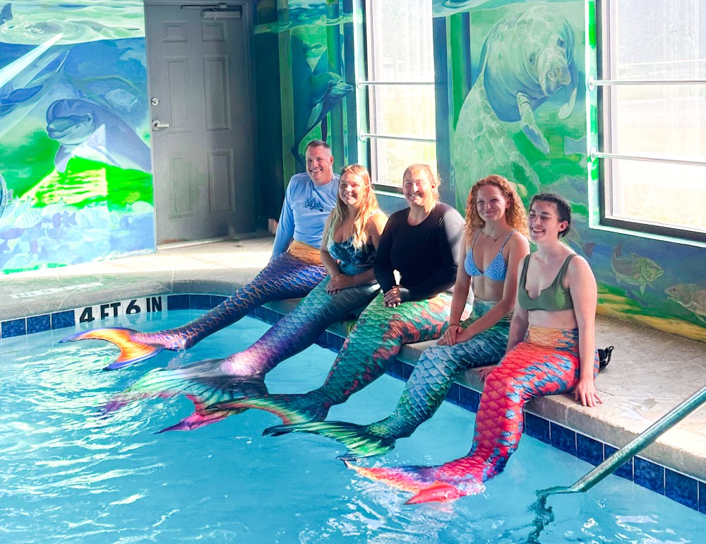 Five people wearing mermaid tails sit by a colorful indoor pool with marine murals.