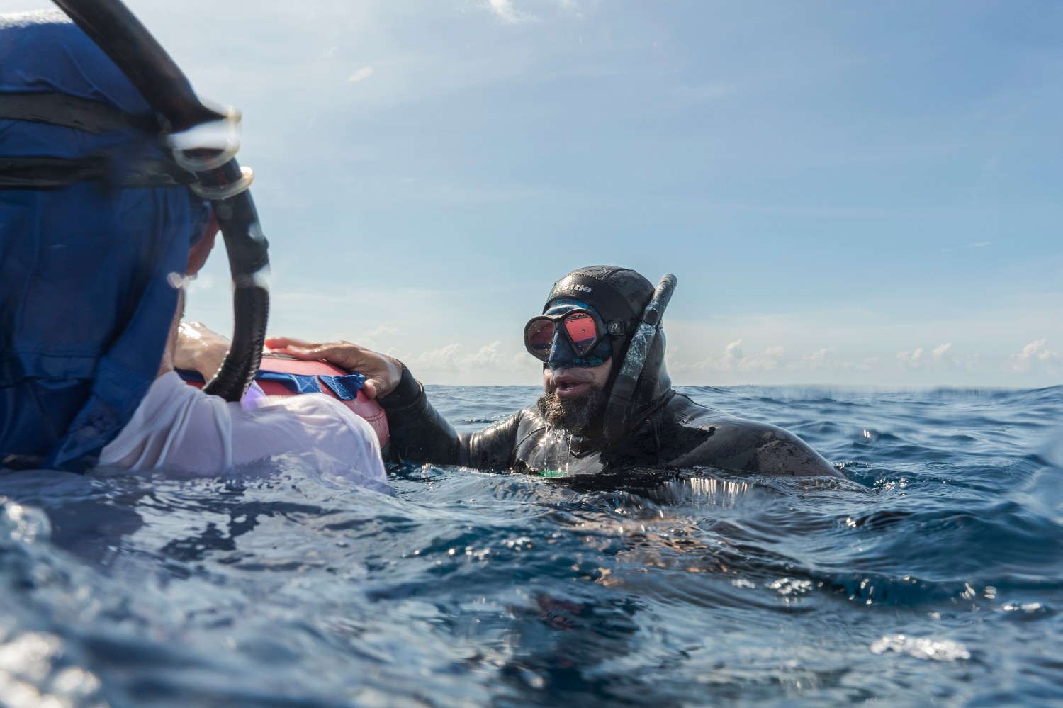 Diver in wetsuit and goggles assisting person in ocean with blue sky background.