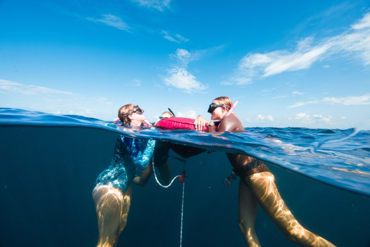 Two snorkelers in clear blue water, sharing a flotation device above and below the surface.