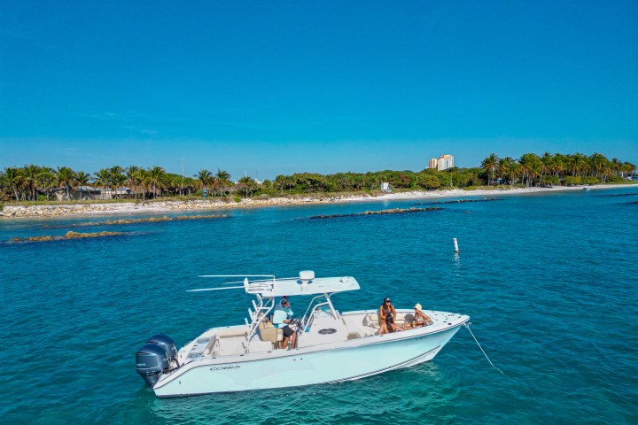 People on a boat in turquoise water near a beach with palm trees and buildings in the background.