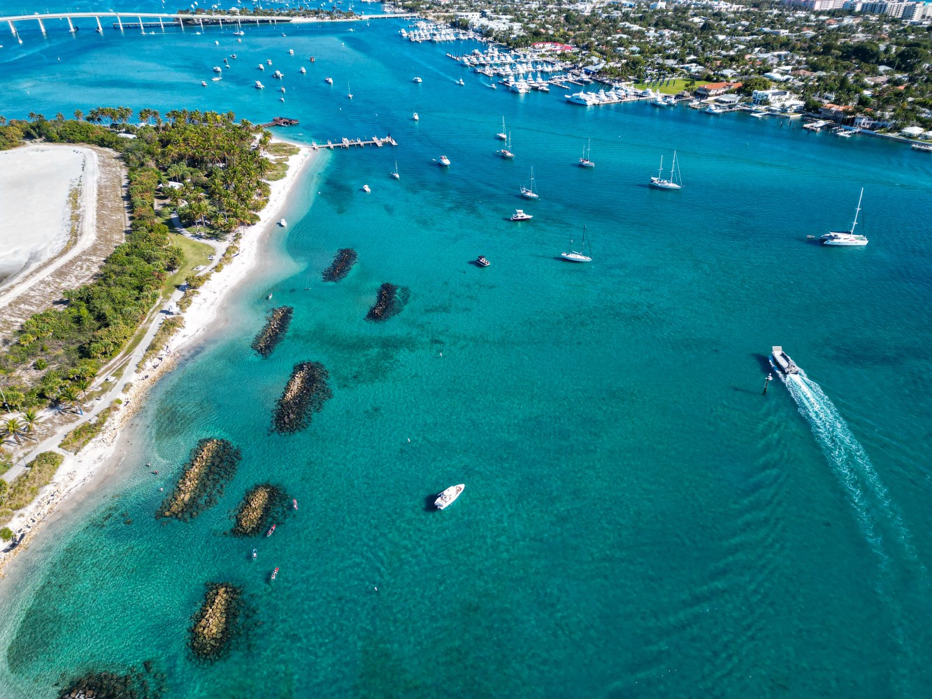 Aerial view of a coastal area with boats, clear blue water, sandy beaches, and green vegetation.