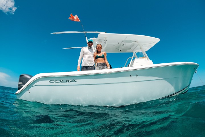 Two people standing on a white boat named COBIA in the ocean under a clear blue sky.