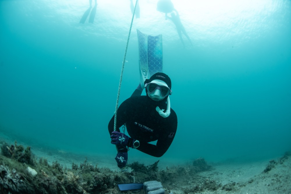 Diver in wetsuit and snorkel underwater holding a rope, with fins and sandy seabed visible.