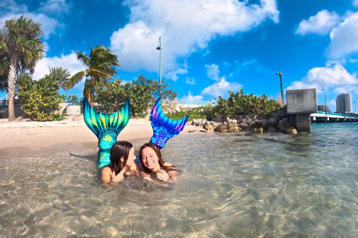Two people with mermaid tails in shallow beach water under a blue sky.