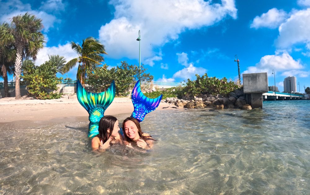 Two people with mermaid tails in shallow beach water under a blue sky.