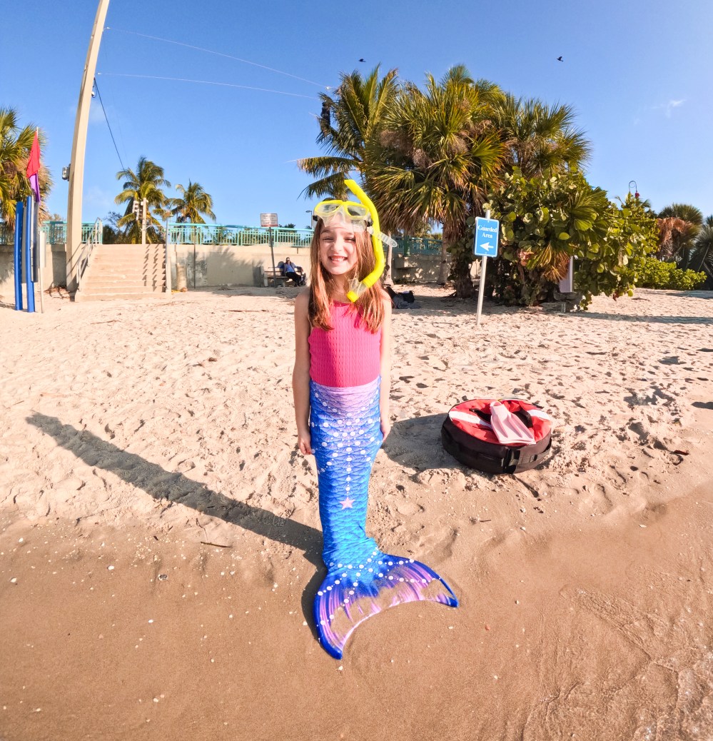 Child in mermaid costume with snorkel stands on sandy beach, palms in background.