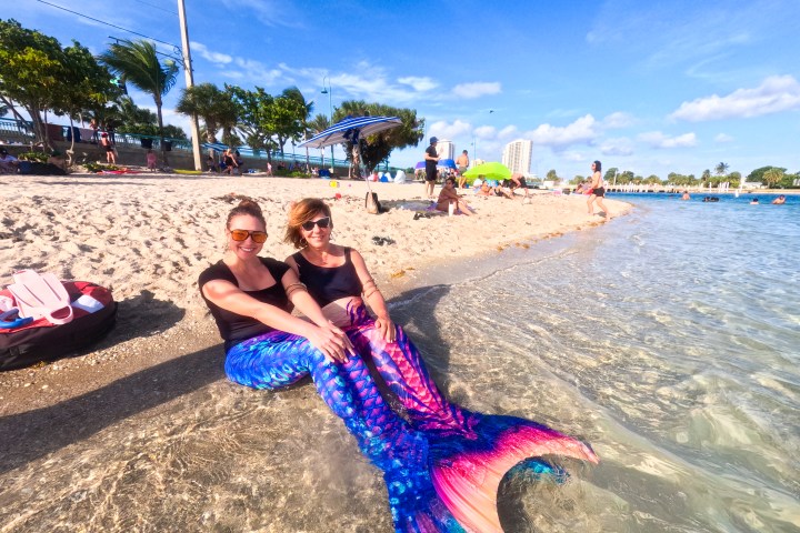 Two women in mermaid costumes sitting by the beach shore with people and umbrellas in the background.
