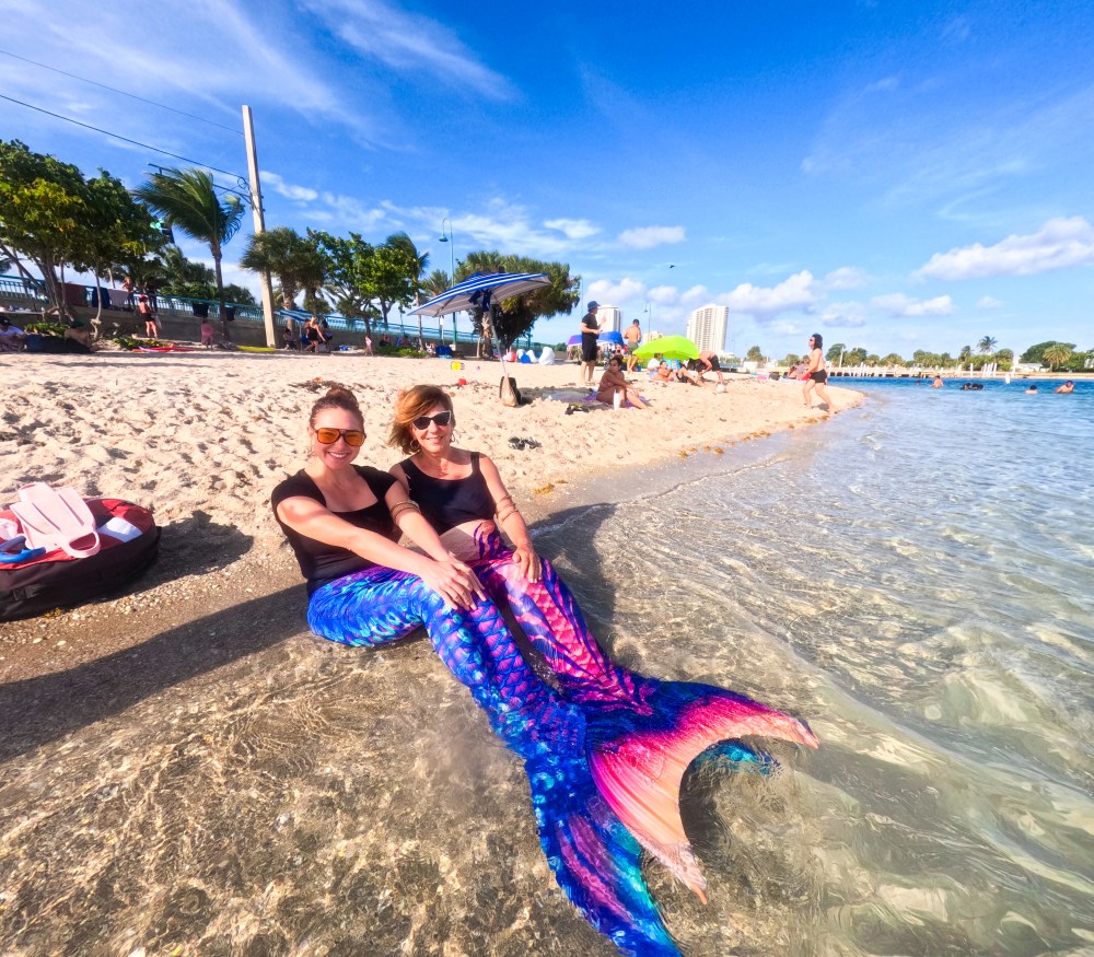 Two women in mermaid costumes sitting by the beach shore with people and umbrellas in the background.