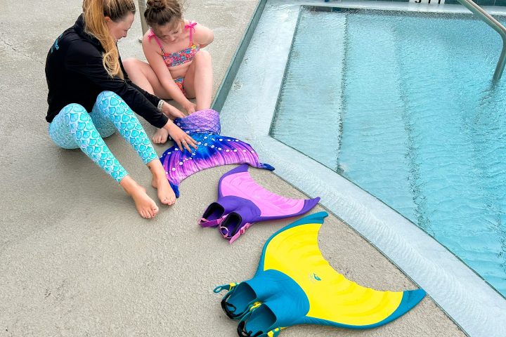 Two people sit by a pool with colorful mermaid tails and fins.