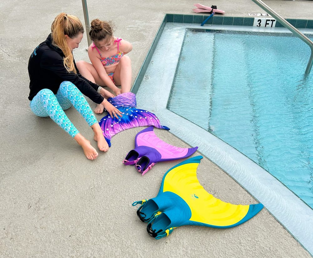 Two people sit by a pool with colorful mermaid tails and fins.