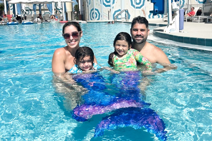 Family in an outdoor pool, two children wearing mermaid tails, with a hotel building in the background.