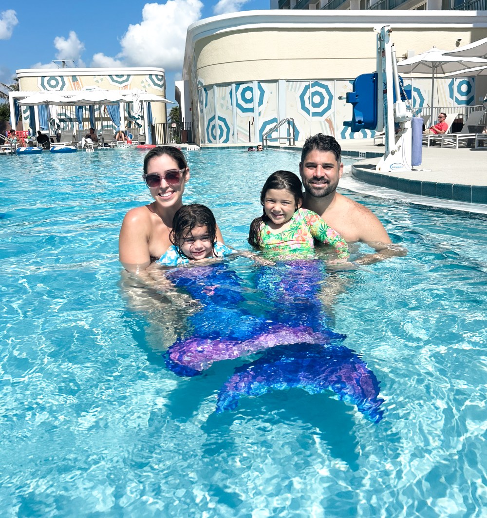 Family in an outdoor pool, two children wearing mermaid tails, with a hotel building in the background.