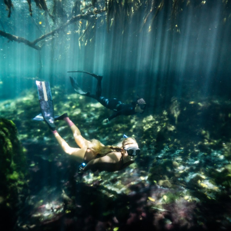 Two divers exploring underwater cave with sunlight streaming through water.