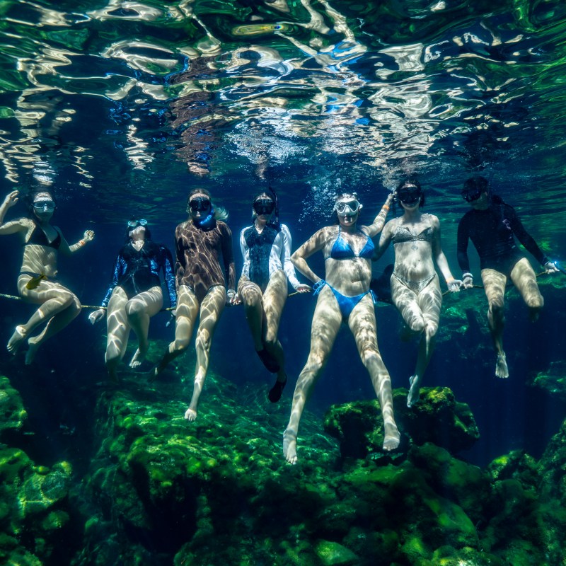 Seven people snorkeling underwater, floating above rocky sea floor.