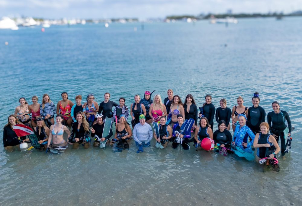 Group of people in swimwear standing in shallow water with a cloudy sky background.