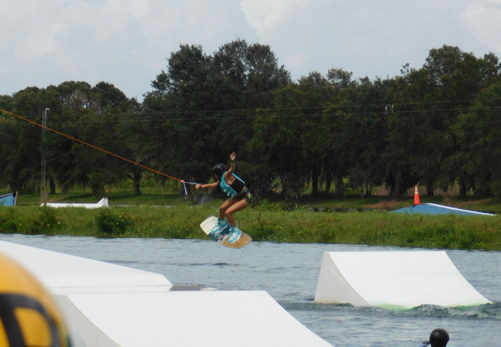 Person wakeboarding on a cable over a water ramp, surrounded by trees and clouds.