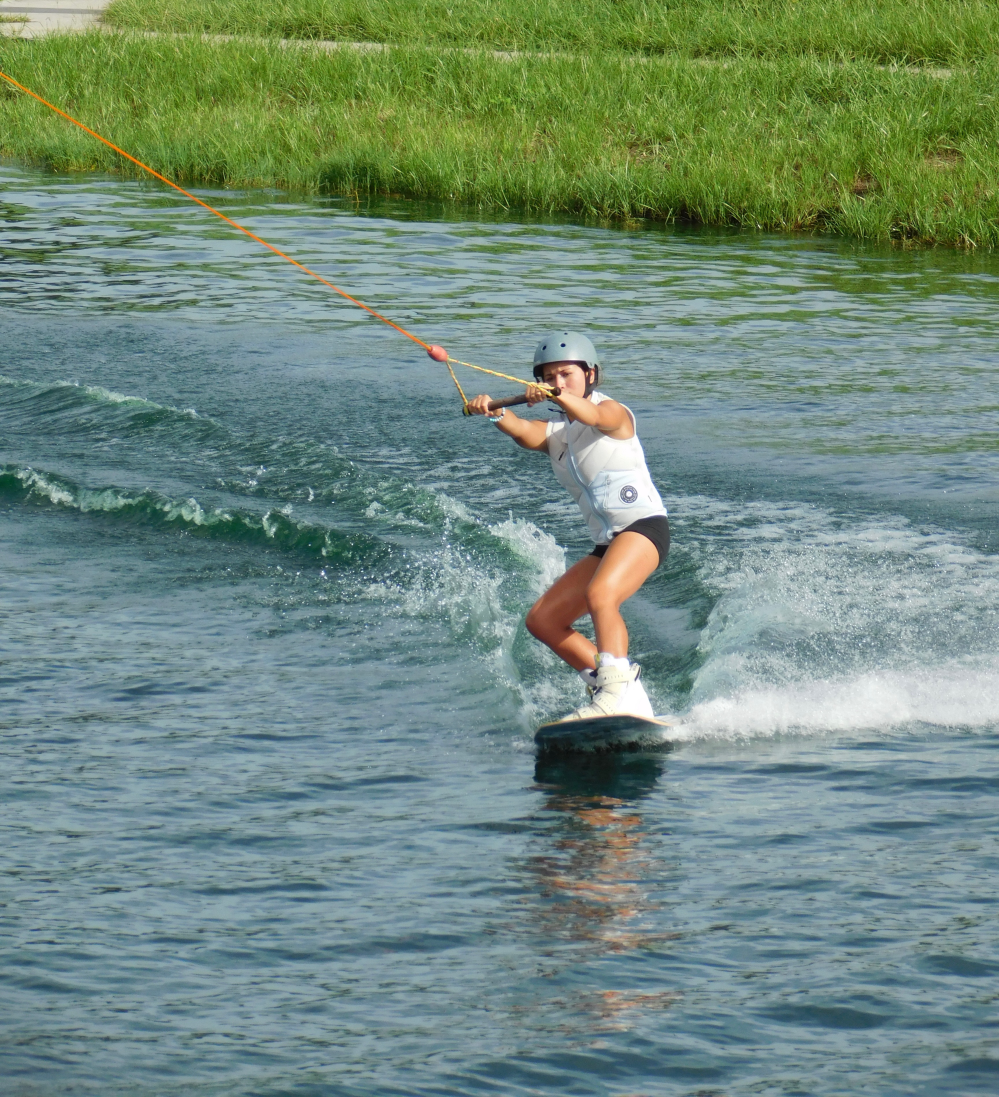 Person wakeboarding on a lake, wearing a helmet and white attire, with a grassy shoreline behind.