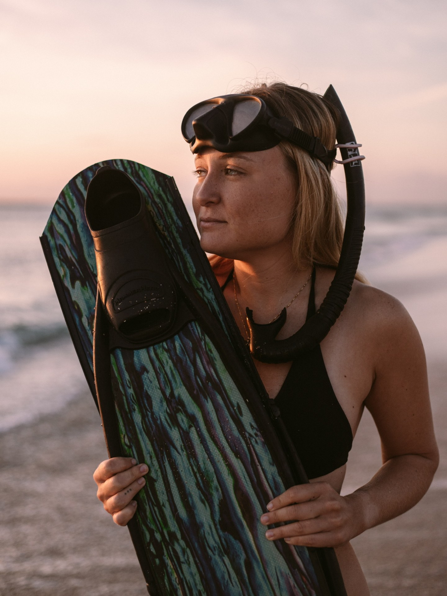 Woman in snorkel gear holding a dive fin on a beach at sunset.