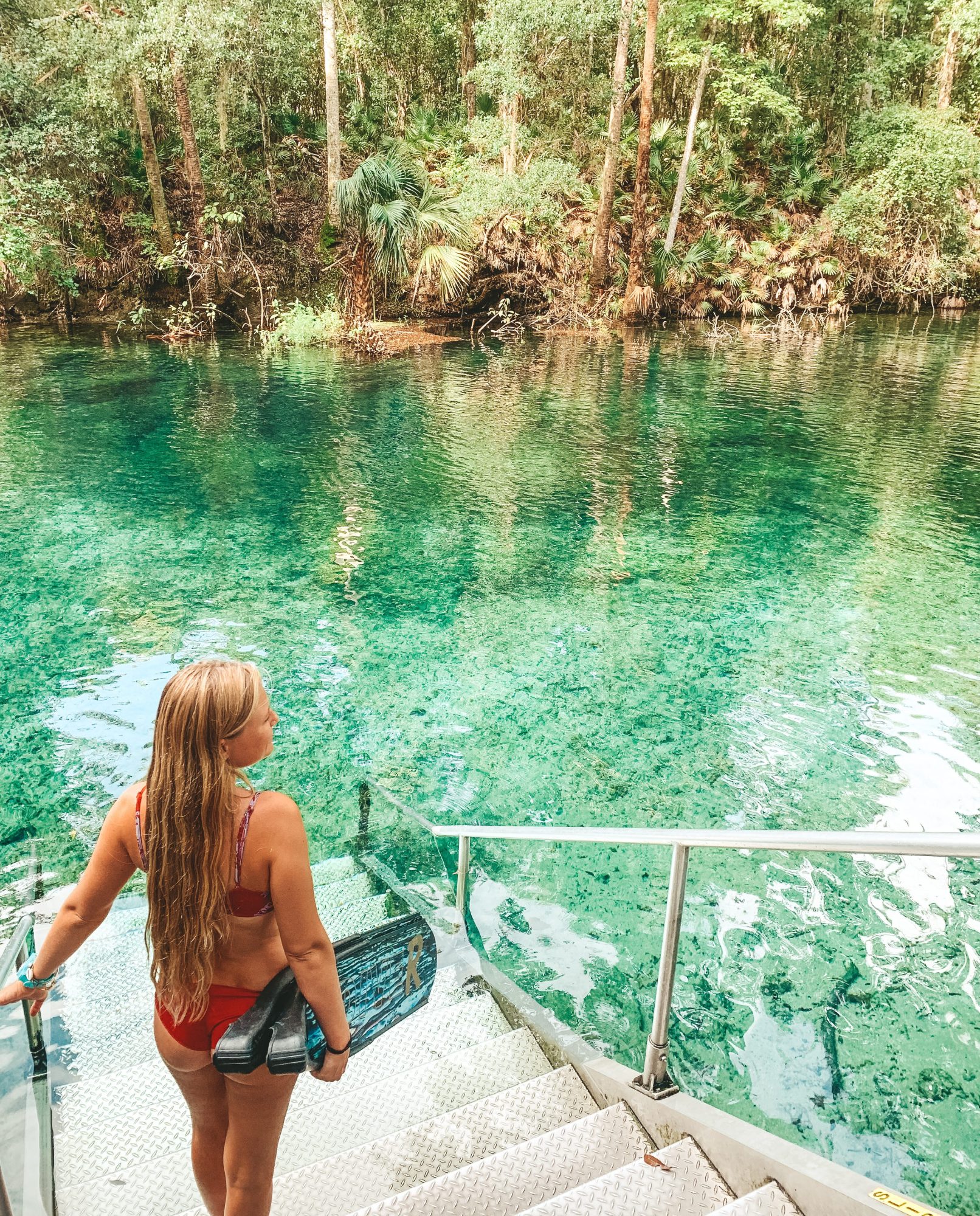 Woman in red bikini holding flippers on stairs by crystal-clear river, surrounded by lush green forest.