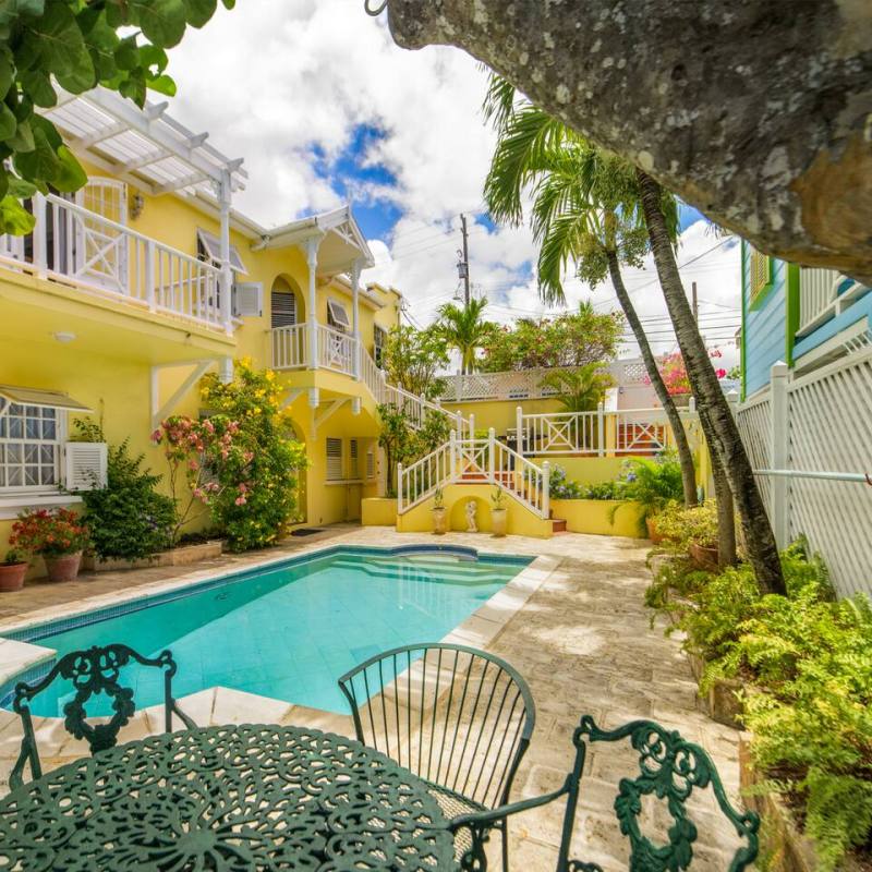 Sunny courtyard with yellow house, pool, green table and chairs, and tropical plants.