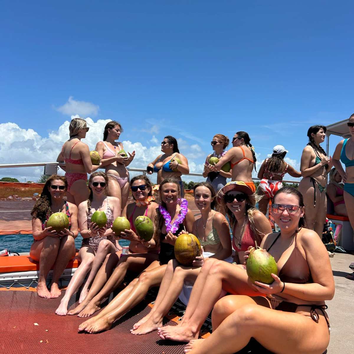 Group of women in swimwear holding coconuts on a boat under a clear sky.