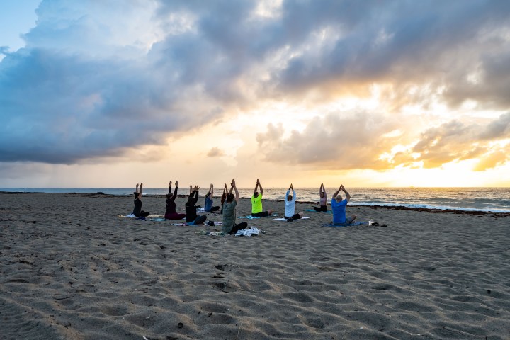 Group practicing yoga on a beach at sunrise with clouds overhead.