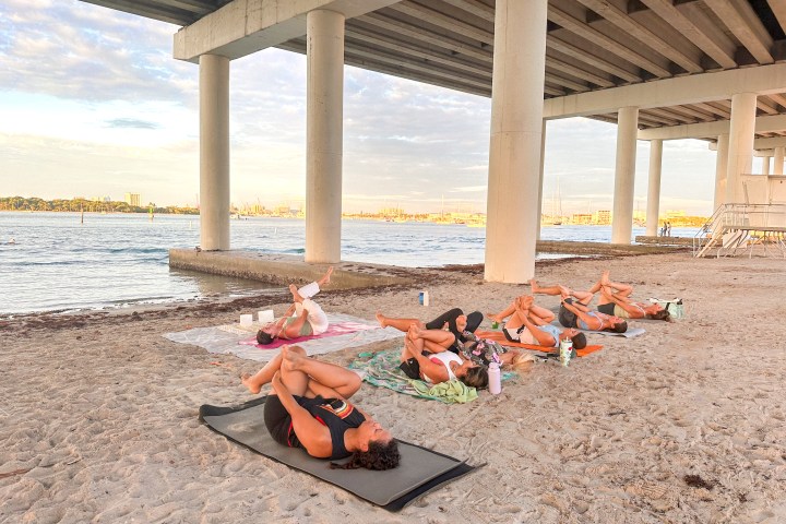 People doing yoga on a sandy beach under Blue Heron Bridge by the water.