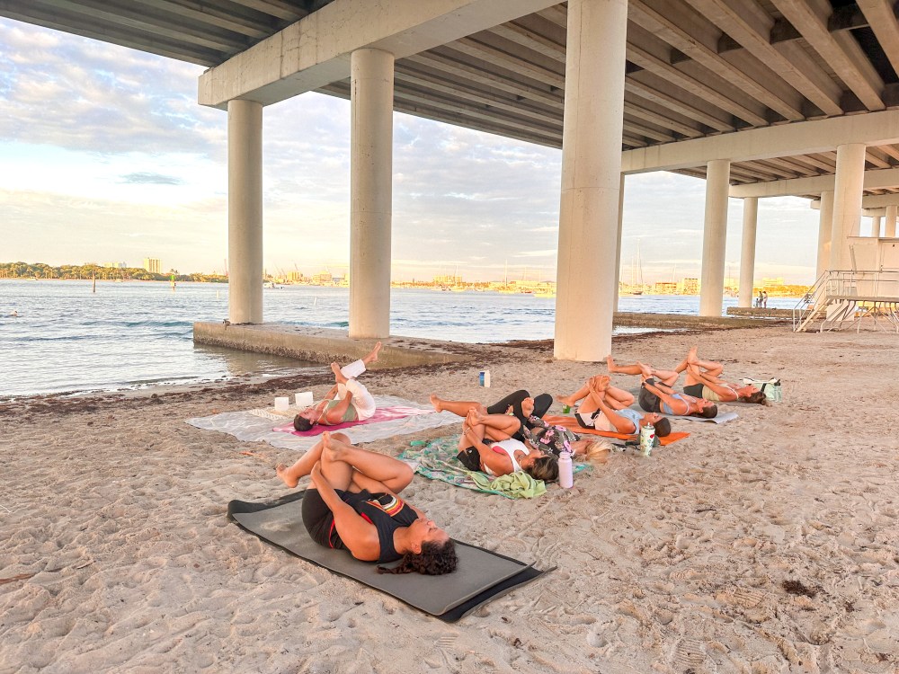 People doing yoga on a sandy beach under Blue Heron Bridge by the water.