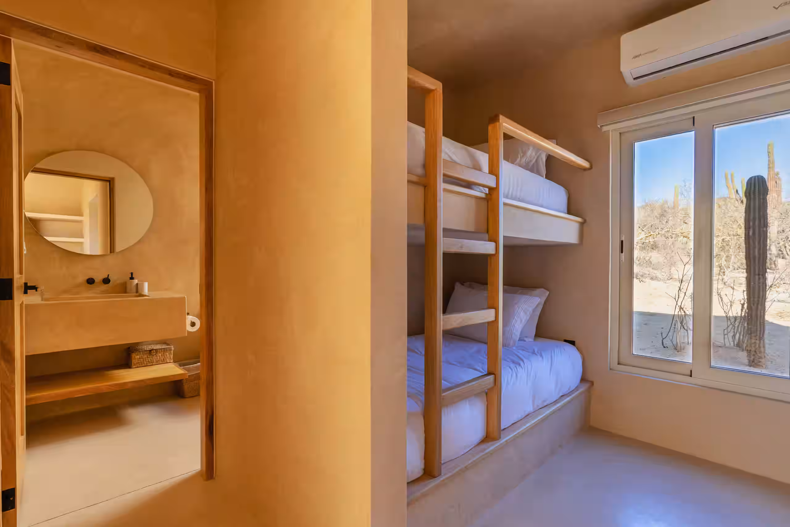 Room with bunk beds, a window showing cacti, and a view into a bathroom with a round mirror.
