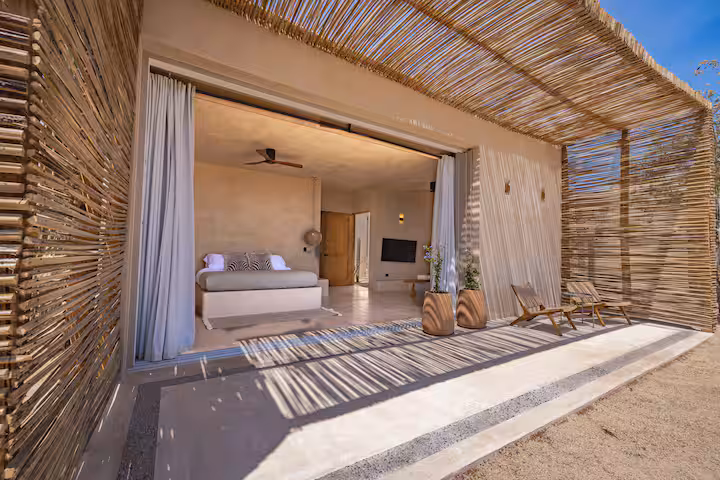 Open-air bamboo patio with a bed, chairs, and curtains under a bright sky.